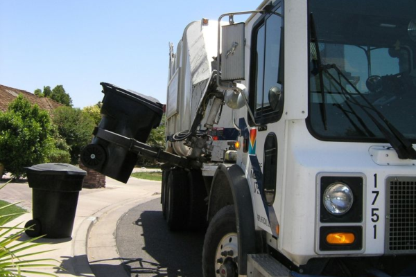 Garbage truck emptying a black trash bin using a mechanical arm on a sunny residential street. Nearby are trees and a brick mailbox.