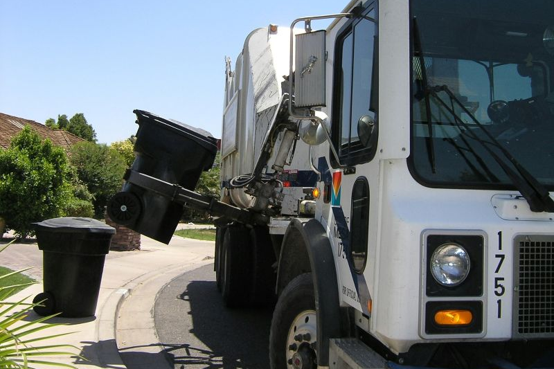 Garbage truck emptying a black trash bin using a mechanical arm on a sunny residential street. Nearby are trees and a brick mailbox.