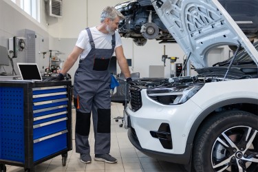 A man in bibs looks under the hood of a white car.