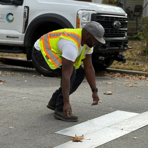 Jonathan "Jay" Young in a fluorescent safety vest crouches beside a white road-marking stencil on pavement, with a work truck behind him.