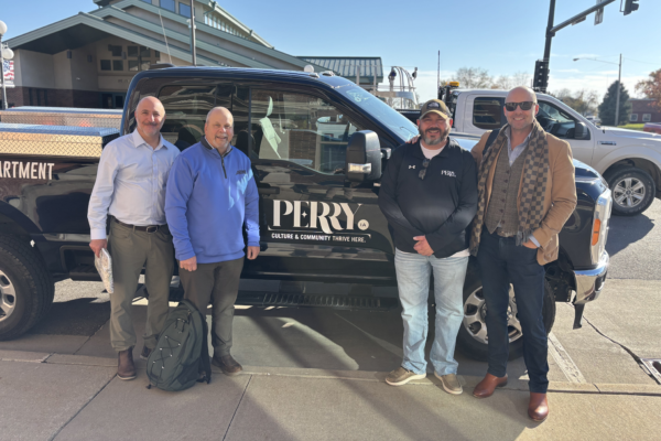 Four men pose beside a black Perry, IA-branded pickup outside a building on a sunny day.