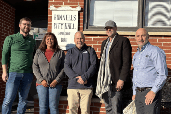 A group of people smiling for a photo. A sign reading "Runnells City Hall Established 1881" hangs behind them.