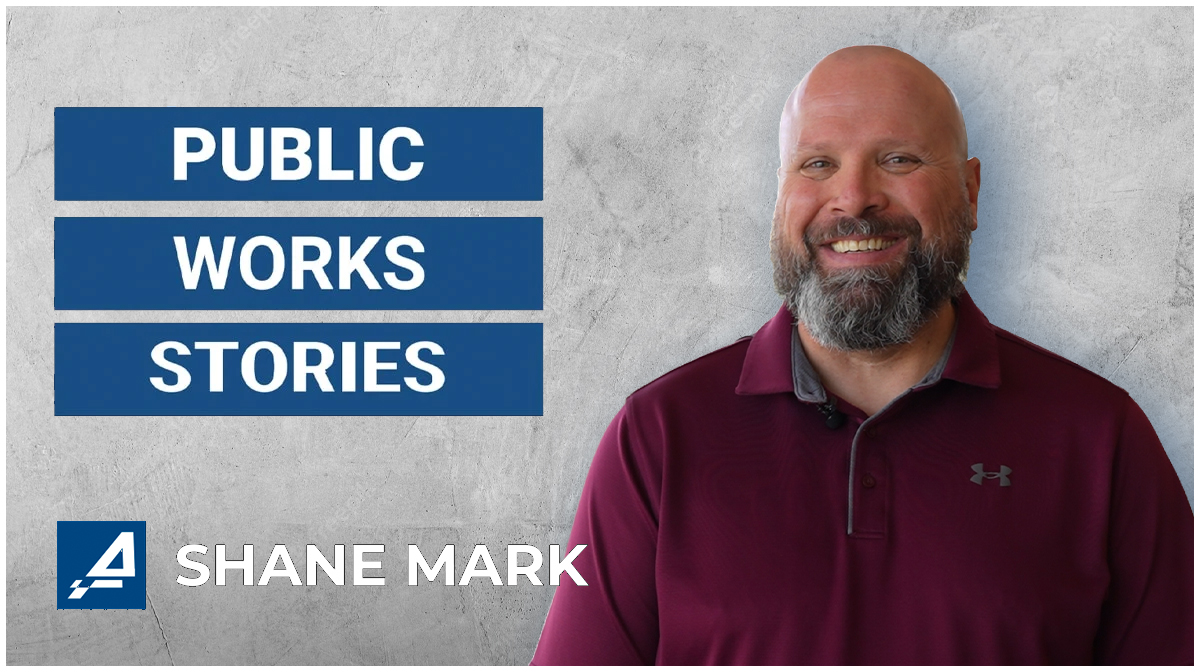 Shane Mark in a maroon shirt stands against a gray background. Blue banners read "Public Works Stories."
