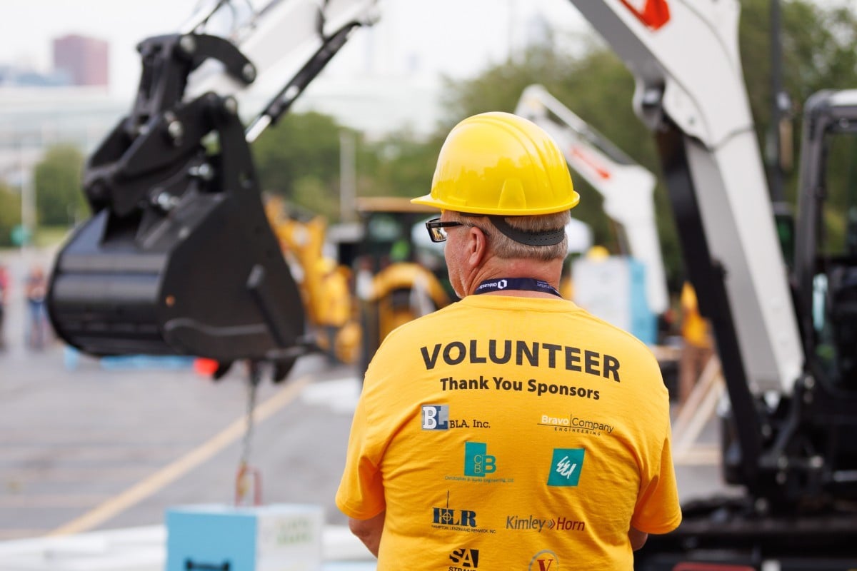 A man in a yellow "volunteer" shirt stands in the foreground while a mini excavator operates in the background.