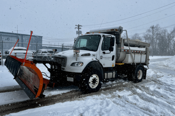 A white truck with plow attached amidst falling snow.