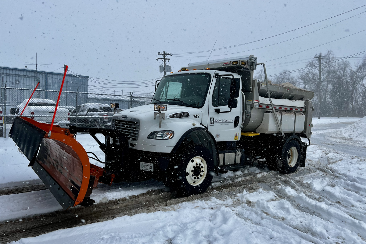 A white truck with plow attached amidst falling snow.
