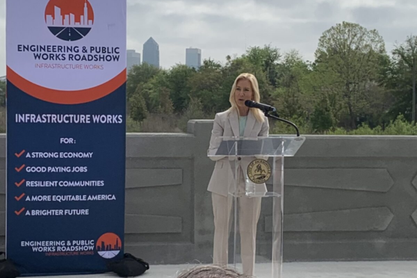A woman in a light suit speaks at a podium outdoors. A banner promoting "Engineering & Public Works Roadshow" is beside her. Trees and cityscape visible.