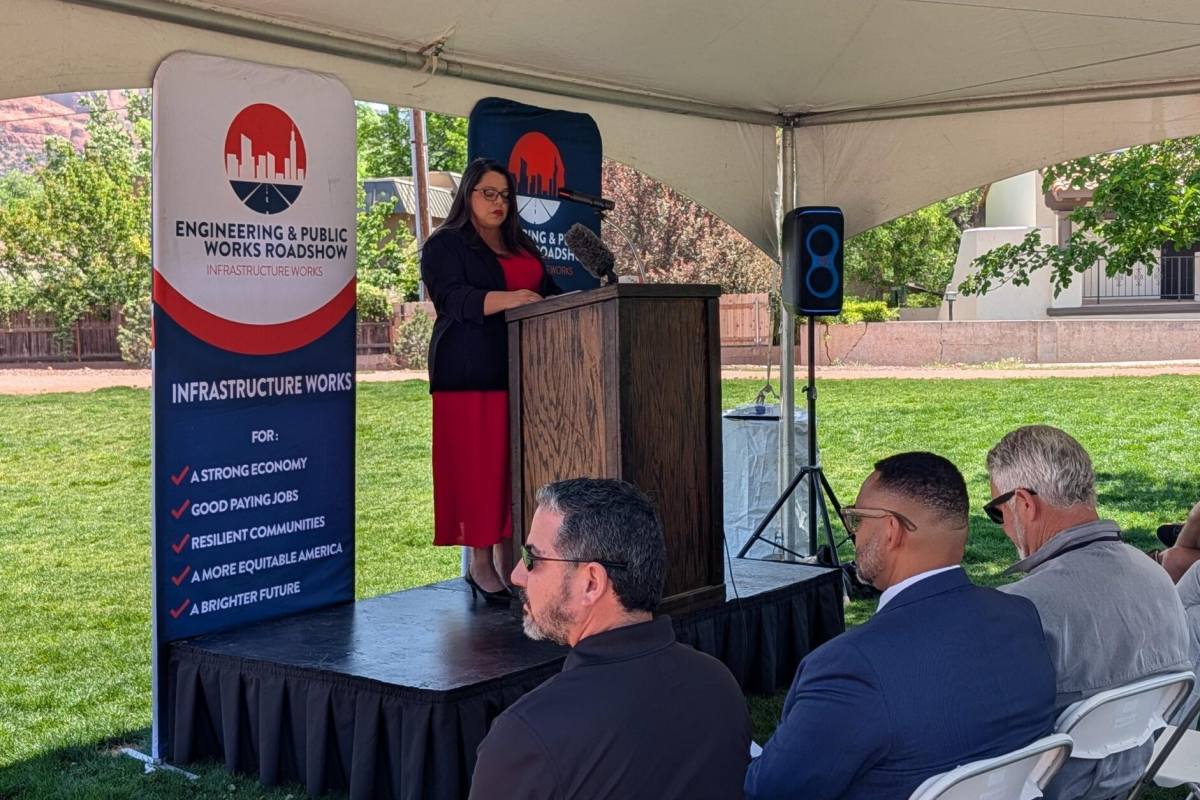 A woman speaks at a lectern under a tent during an outdoor event. A banner beside her reads "Engineering & Public Works Roadshow." Attendees sit and listen attentively.