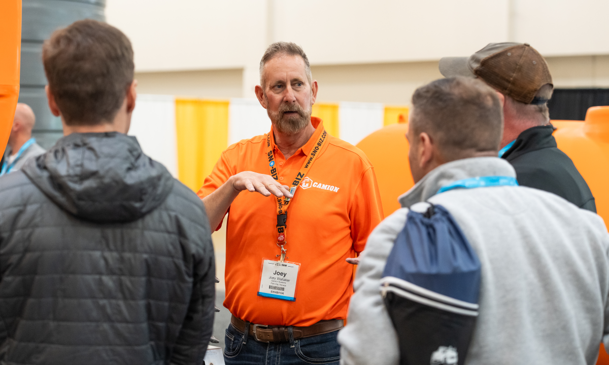 An exhibitor in an orange polo shirt discusses his company's products with attendees at the APWA Snow Conference.