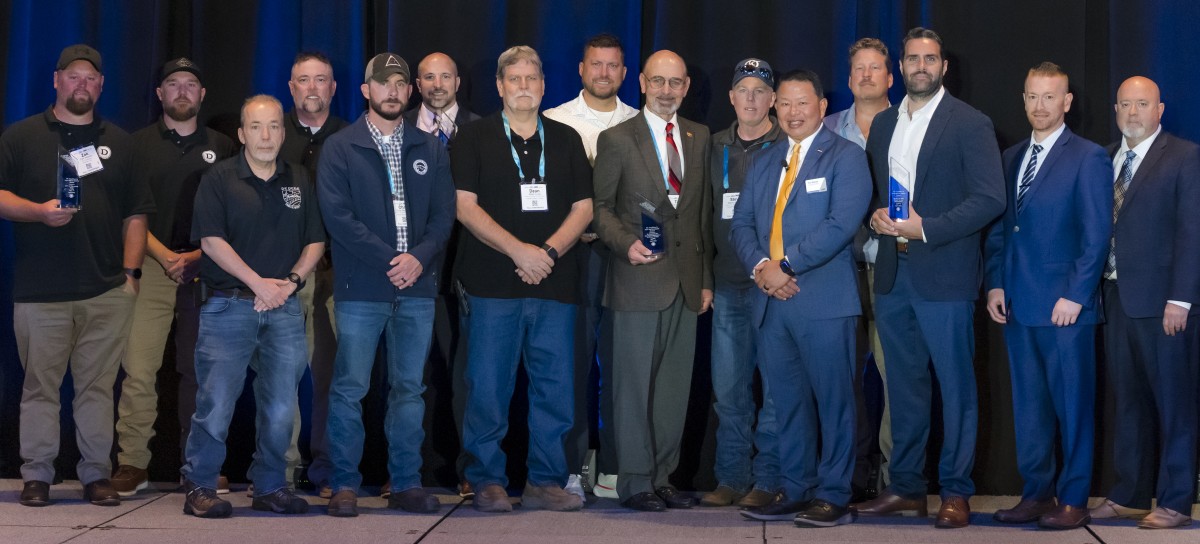 A group of 15 men stands side by side on a stage against a dark curtain, dressed in a mix of casual and formal attire, holding awards and smiling.