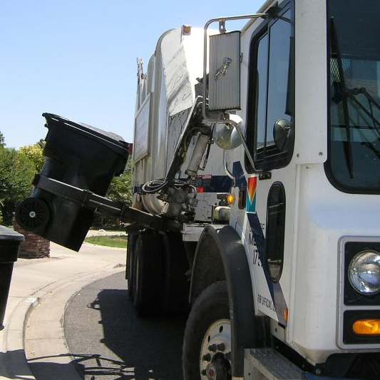 Garbage truck emptying a black trash bin using a mechanical arm on a sunny residential street. Nearby are trees and a brick mailbox.