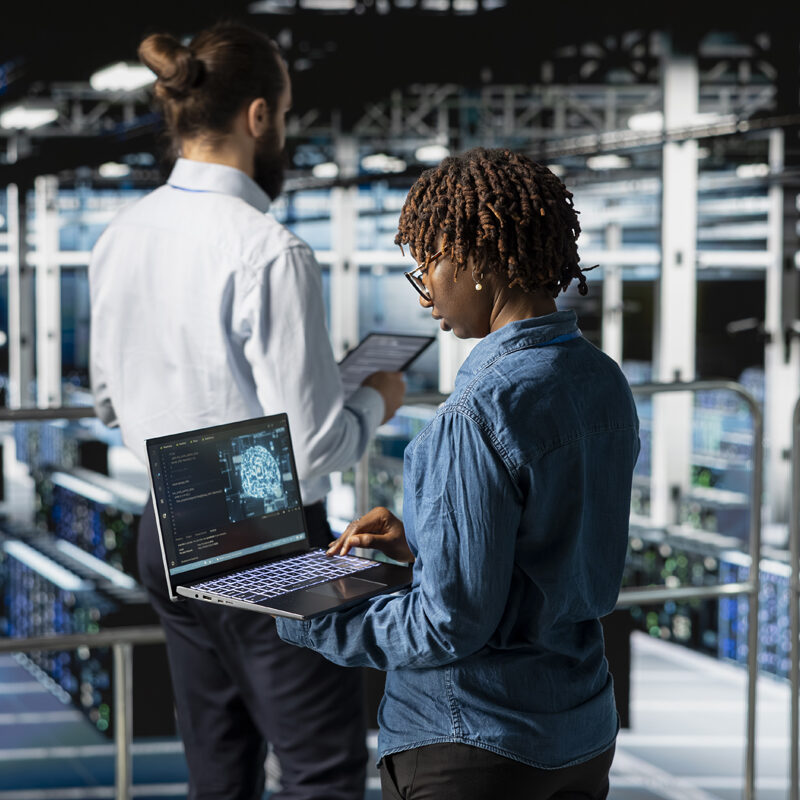 Two individuals stand in a spacious data center, each using an electronic device.