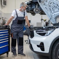 A man in bibs looks under the hood of a white car.