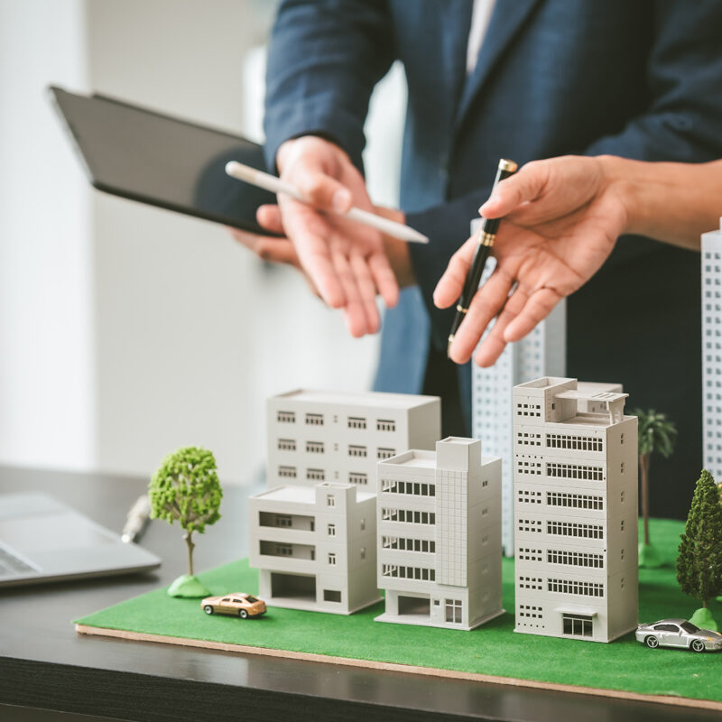 Two middle-aged Asian people businessmen, one holding wind turbine model, discussing over model city, likely about clean energy and new housing projects.