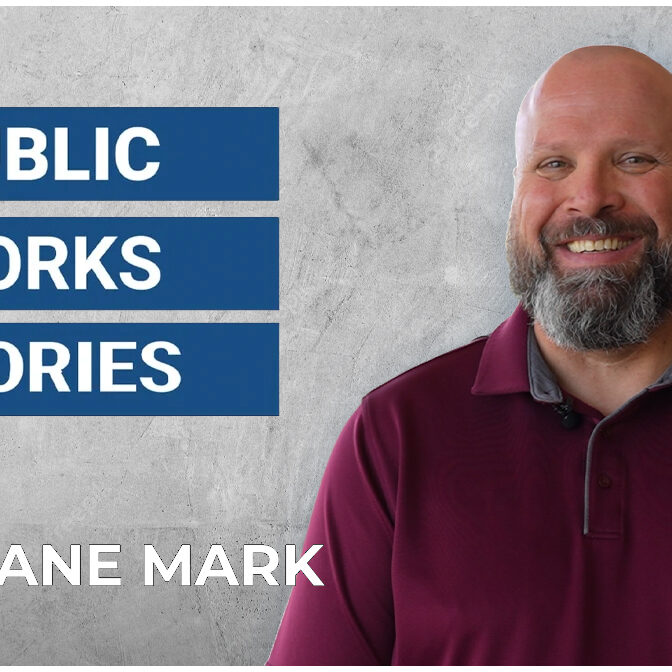 Shane Mark in a maroon shirt stands against a gray background. Blue banners read "Public Works Stories."