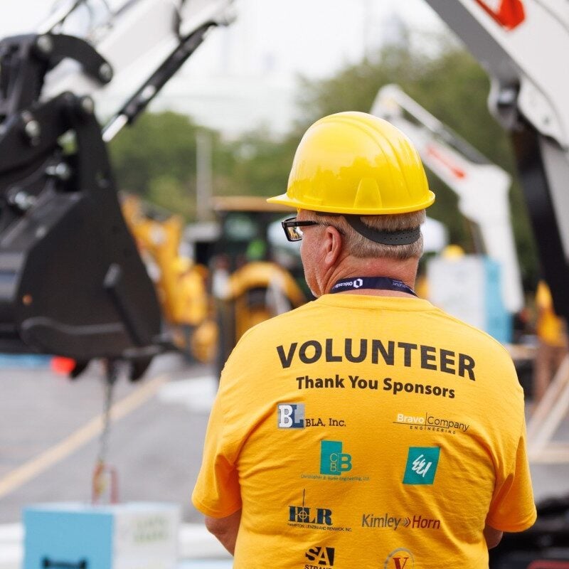 A man in a yellow "volunteer" shirt stands in the foreground while a mini excavator operates in the background.