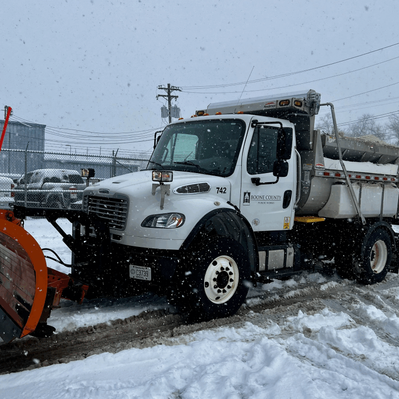 A white truck with plow attached amidst falling snow.