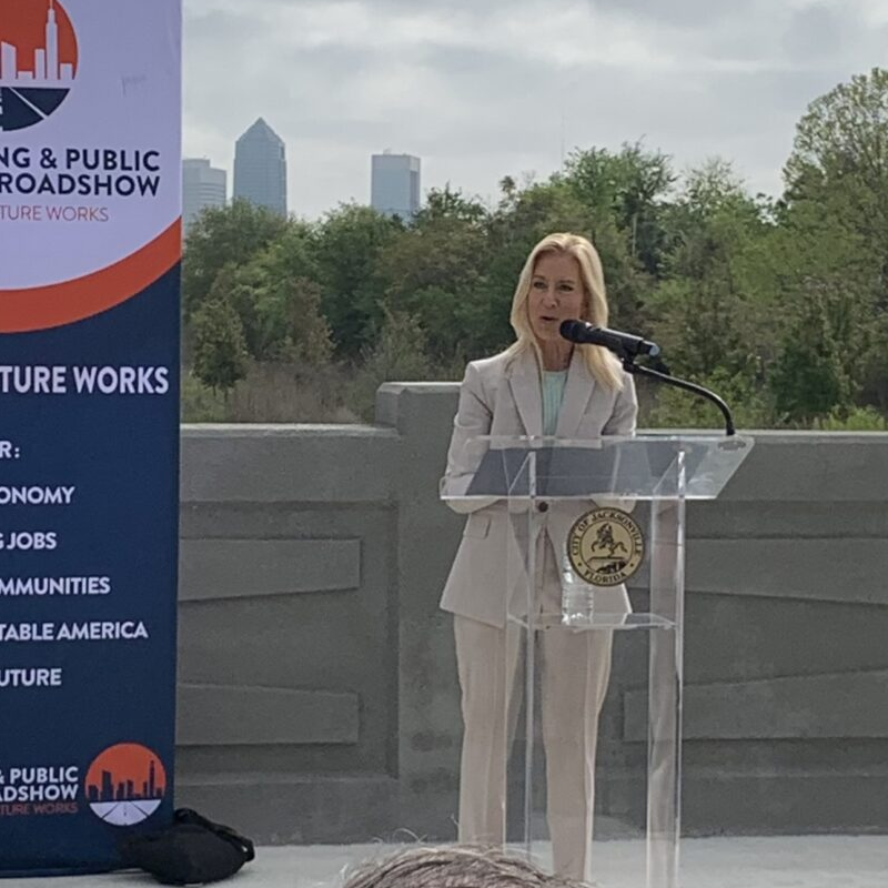A woman in a light suit speaks at a podium outdoors. A banner promoting "Engineering & Public Works Roadshow" is beside her. Trees and cityscape visible.