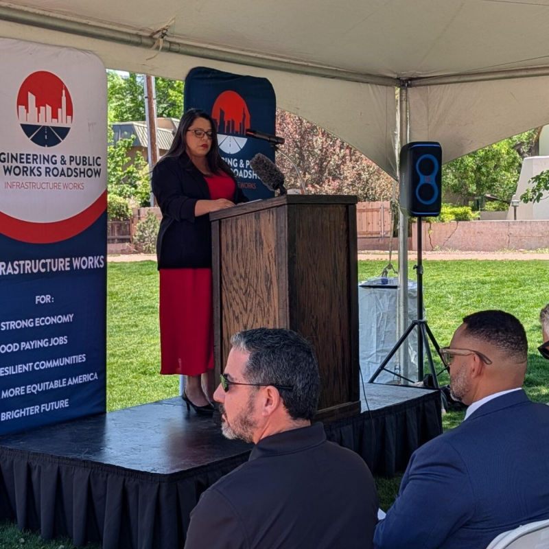 A woman speaks at a lectern under a tent during an outdoor event. A banner beside her reads "Engineering & Public Works Roadshow." Attendees sit and listen attentively.